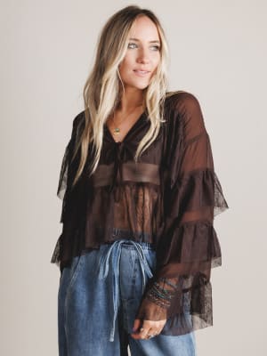 Woman wearing a sheer brown blouse with ruffled sleeves and blue jeans against a plain background