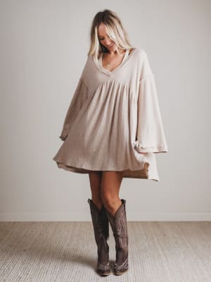 Woman wearing a beige dress and brown boots in a room with a plain background