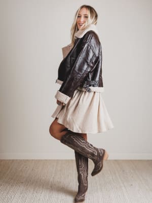 Person wearing a leather jacket, beige dress, and brown boots in a minimal indoor setting.