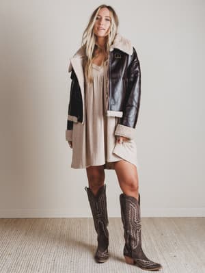 Woman wearing a black leather jacket with shearling trim, a beige dress, and brown cowboy boots indoors.