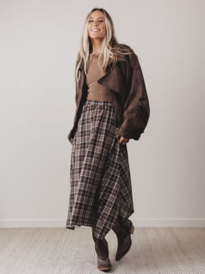 Woman wearing a brown coat, brown top, and plaid skirt against a plain background
