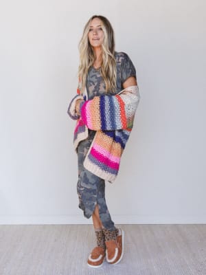 Woman wearing a long camouflage dress with patterned socks and loafer mules, along with a brightly colored oversized cardigan.