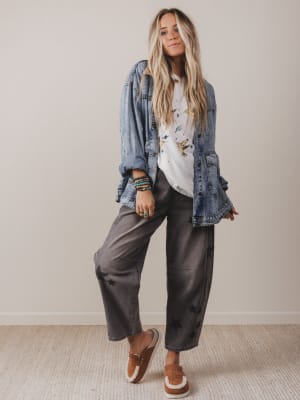 Woman wearing a denim jacket, white shirt, and gray pants against a plain background