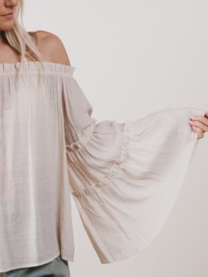 Woman wearing a light-colored off-shoulder blouse with flowing sleeves against a plain background