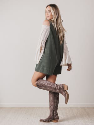 Woman wearing a green dress, white blouse, and brown knee-high boots against a plain background