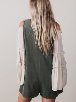 Person wearing a green romper with a white blouse underneath on a plain background