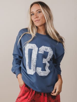 Woman wearing a blue sweatshirt with '03' printed on it against a beige background