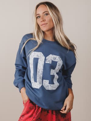Woman wearing a blue sweatshirt with '03' printed on it against a beige background