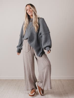 Woman wearing a gray sweater and patterned pants standing against a plain background