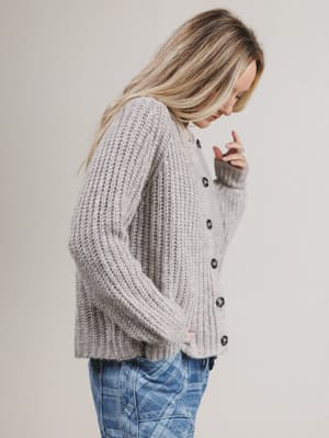 Side view of woman wearing a chunky knit cardigan and jeans against a plain background