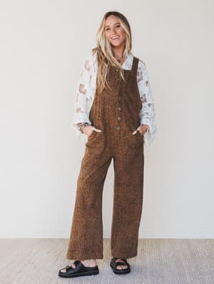 Woman wearing a brown patterned jumpsuit with a white lace top underneath, standing against a plain background.