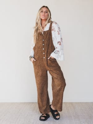 Woman wearing a brown patterned jumpsuit with a white lace top against a plain background
