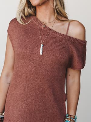 A rust-toned knit sweater dress styled off one shoulder, layered with delicate gold necklaces and stacked beaded bracelets — the kind of effortless outfit that looks like it came together naturally but definitely didn't.