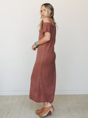 A rust knit midi dress with an off-shoulder neckline worn with tan wooden clogs and a stack of turquoise bracelets — effortless and a little earthy, like she's headed somewhere worth going.