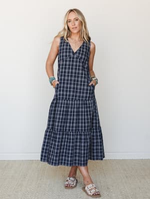 A navy plaid midi dress with a relaxed tiered skirt and hands-in-pockets ease — styled with stacked boho bracelets and sandals against a clean studio wall, the kind of dress that works just as hard at brunch as it does running around all day.