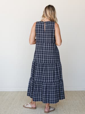 A tiered maxi dress in navy and white plaid photographed from behind, showing a relaxed silhouette with a keyhole back detail and neutral slide sandals — the kind of dress that works just as hard at brunch as it does at a summer farmers market.