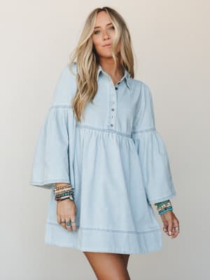 A light denim mini dress with dramatic bell sleeves and a babydoll silhouette, styled with stacks of boho bracelets and rings against a soft neutral backdrop — effortless and a little free-spirited.