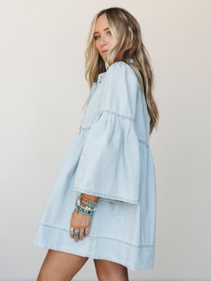 A light denim mini dress with a relaxed, oversized silhouette shot in profile — the bell sleeves and tiered seaming give it an easy boho feel, styled with a stack of turquoise and gold bracelets and rings that feel collected, not coordinated.