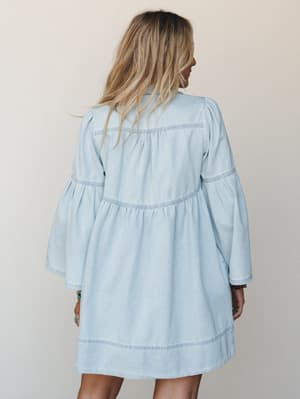 A breezy light denim mini dress shot from the back, with voluminous bell sleeves, tiered smocking details, and a relaxed babydoll silhouette — the kind of dress that looks effortless whether you're running errands or catching a set at an outdoor show