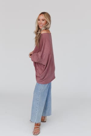 A woman with long blonde hair smiles while standing sideways in The Nest Sunday Stroll Oversized Tee - Mauve, paired with light blue wide-leg jeans and brown sandals against a plain white background.