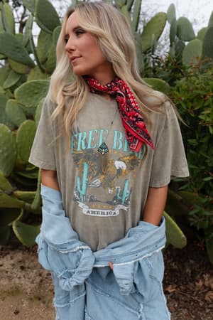 A woman with long blonde hair stands before cacti, wearing the oversized Fly Free Graphic Tee - Mocha for a boho-chic vibe, paired with a red bandana and a denim jacket draped at her waist as she looks to the side.