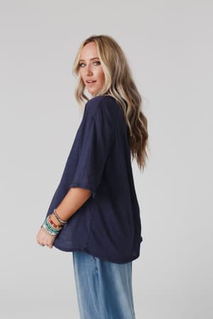 A woman with long, wavy blonde hair models The Nest Your Favorite Slub Tee Shirt in Navy and light blue jeans, accessorized with colorful bracelets. She stands against a plain light background, glancing over her shoulder.