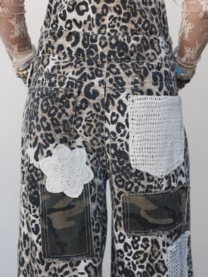 A close-up back view of leopard print overalls layered with camo patches and crochet pocket details, styled with a sheer floral lace long-sleeve underneath — the kind of outfit that commits fully and doesn't apologize for it.