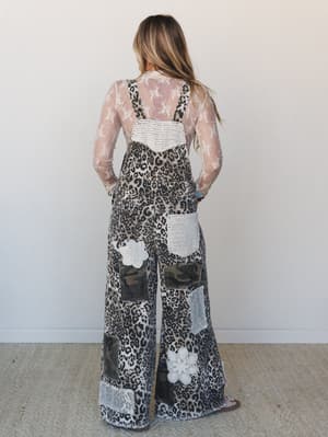 Shot from the back, a model wears wide-leg leopard print overalls layered over a sheer lace long-sleeve top — the patchwork mix of camo panels, crochet patches, and floral appliqués gives the whole look a collected, one-of-a-kind energy.