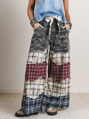 Person wearing a patchwork pants with camouflage, plaid, and checkered patterns on a neutral background.