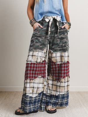 Person wearing a patchwork pants with camouflage, plaid, and checkered patterns on a neutral background.