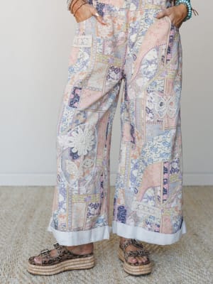 Wide-leg patchwork print pants with crochet appliqué details — a butterfly on one leg, a daisy on the other — styled with stacked bracelets and leopard print platform sandals against a soft neutral interior backdrop.