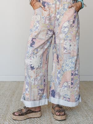 Wide-leg patchwork print pants with crochet appliqué details — a butterfly on one leg, a daisy on the other — styled with stacked bracelets and leopard print platform sandals against a soft neutral interior backdrop.