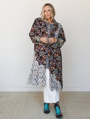 A layered boho look built around a long paisley-and-floral patchwork duster with sheer lace panels, worn open over a gingham top and wide-leg white jeans, finished with turquoise cowboy boots and stacked silver jewelry.