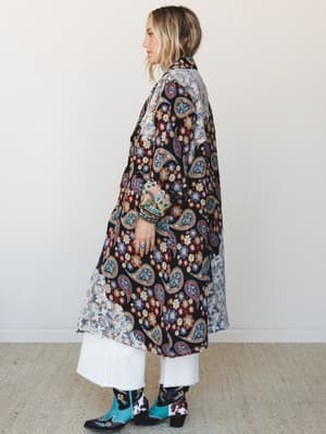 A model shot in profile wearing a dramatic mixed-print duster jacket over white wide-leg jeans and turquoise cowboy boots — the kind of outfit that makes people stop you in a parking lot to ask where you got it.