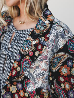 A close-up detail shot of a richly patterned jacket layered over a gingham top — mixed prints, mismatched decorative buttons, and sheer embroidered overlay give it that collected, found-at-a-market feel.