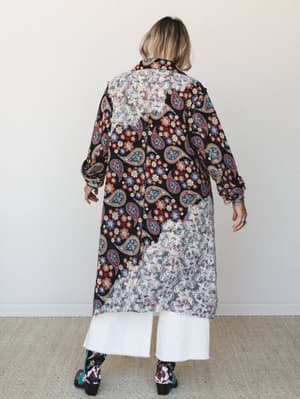 Shot from the back, a model wears a dramatic long printed duster over wide-leg white jeans and printed boots — the mixed paisley and sheer lace-panel fabric gives it that perfectly collected, lived-in boho energy.