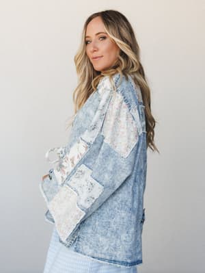A model looks over her shoulder in a roomy acid-wash denim jacket covered in patchwork floral fabric panels — the kind of piece that feels like it was thrifted, then treasured.