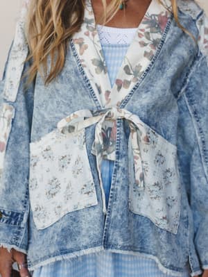 A close-up of a mixed-media denim jacket with floral patchwork details and a front tie closure, layered over a light blue gingham dress — the kind of piece that looks like it was thrifted, loved, and styled intentionally.