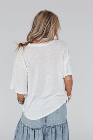 A woman with wavy blonde hair stands facing away, wearing The Nest Your Favorite Slub Tee Shirt in Natural and a light blue tiered denim skirt, accessorized with colorful bracelets, against a plain light background.