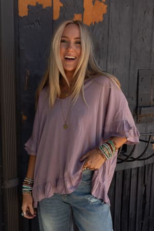 A smiling woman with long blonde hair wears The Nest Let Me Be Ruffled Top in Pale Plum, featuring a lightweight stretch knit and v-neckline. She pairs it with ripped jeans and layered necklaces, standing against a textured dark wall.