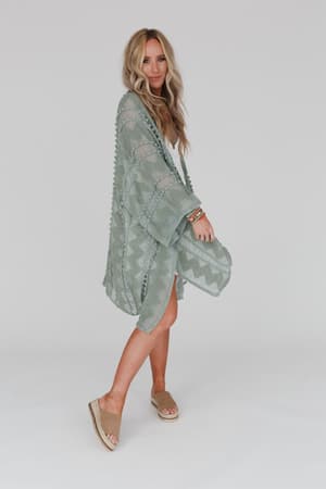 A woman with long blonde hair wears the Bali Embroidered Kimono - Sage, a light green boho piece over a white dress, paired with tan slide sandals and bracelets, standing against a plain white background.