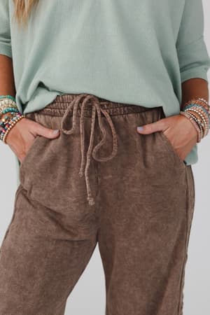 A person wears a light green long-sleeve top and So Comfy Wide Leg Pant Full Length - Mocha, with hands in pockets, showcasing multiple colorful beaded bracelets on both wrists.
