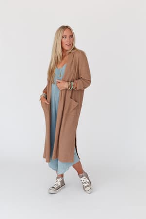 A woman with long blonde hair wears a light blue dress, bracelets, gray high-top sneakers, and The Nest Jayde Waffle Knit Cardigan in Taupe, standing against a plain white background.