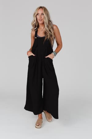 A woman with long blonde hair wears The Nest Serenity Wide Leg Jumpsuit in black, styled with beige sandals and colorful bracelets. She stands with her hands in her pockets against a plain light gray background.