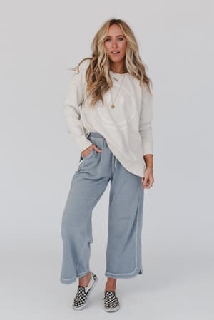 A woman with long blonde hair wears a white long-sleeve shirt, the So Comfy Wide Leg Pant Cropped Length in Faded Blue, and black-and-white checkerboard slip-on shoes, standing against a plain white background.
