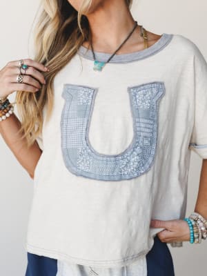 A relaxed cream tee with a patchwork horseshoe appliqué in dusty blue takes center stage, styled with stacked boho bracelets, silver rings, and a turquoise pendant — the kind of top that feels effortless and a little sentimental at the same time.