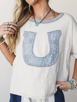 A relaxed cream tee with a patchwork horseshoe appliqué in dusty blue takes center stage, styled with stacked boho bracelets, silver rings, and a turquoise pendant — the kind of top that feels effortless and a little sentimental at the same time.