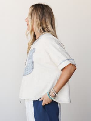 A relaxed, side-profile shot of a cream oversized tee with a dusty blue embroidered bib-style chest panel and rolled sleeves, styled with navy wide-leg pants and a stack of beaded bracelets — the kind of effortless outfit that looks like it came toge