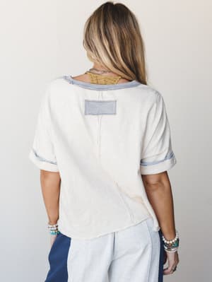 A back-facing shot of a relaxed cream tee with contrast periwinkle trim and a visible patch detail — styled with wide-leg pants and stacked bracelets for that effortless, lived-in feel.