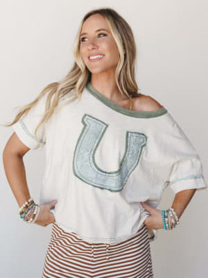 A relaxed, off-shoulder graphic tee in cream with a sage green horseshoe appliqué patch takes center stage, styled with rust-striped bottoms and stacked boho bracelets — the kind of effortless outfit that feels as good as it looks.